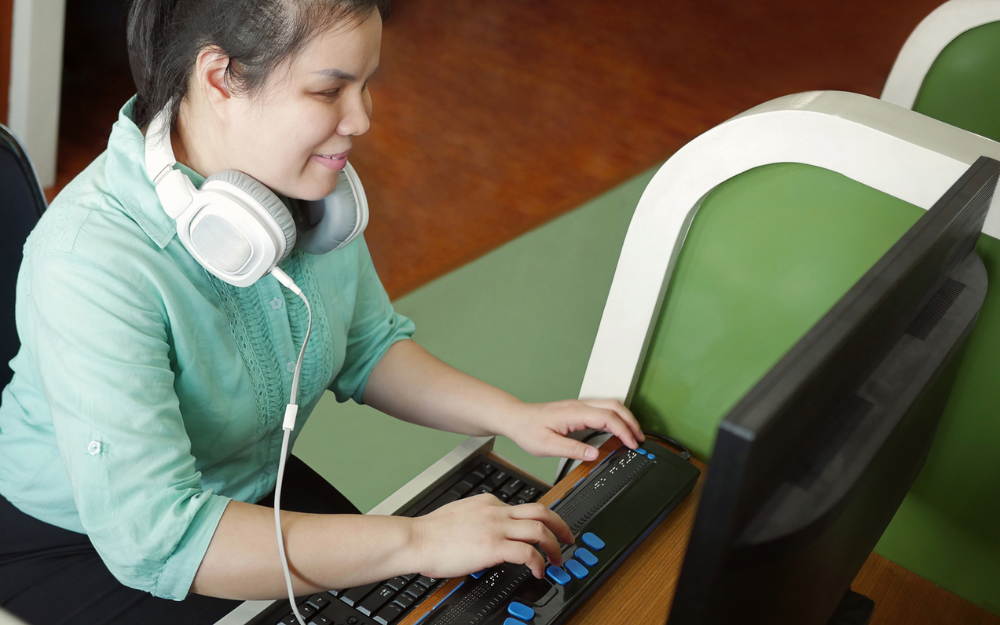 young blind woman with headphone using computer with refreshable braille display or braille terminal a technology device for persons with visual disabilities.